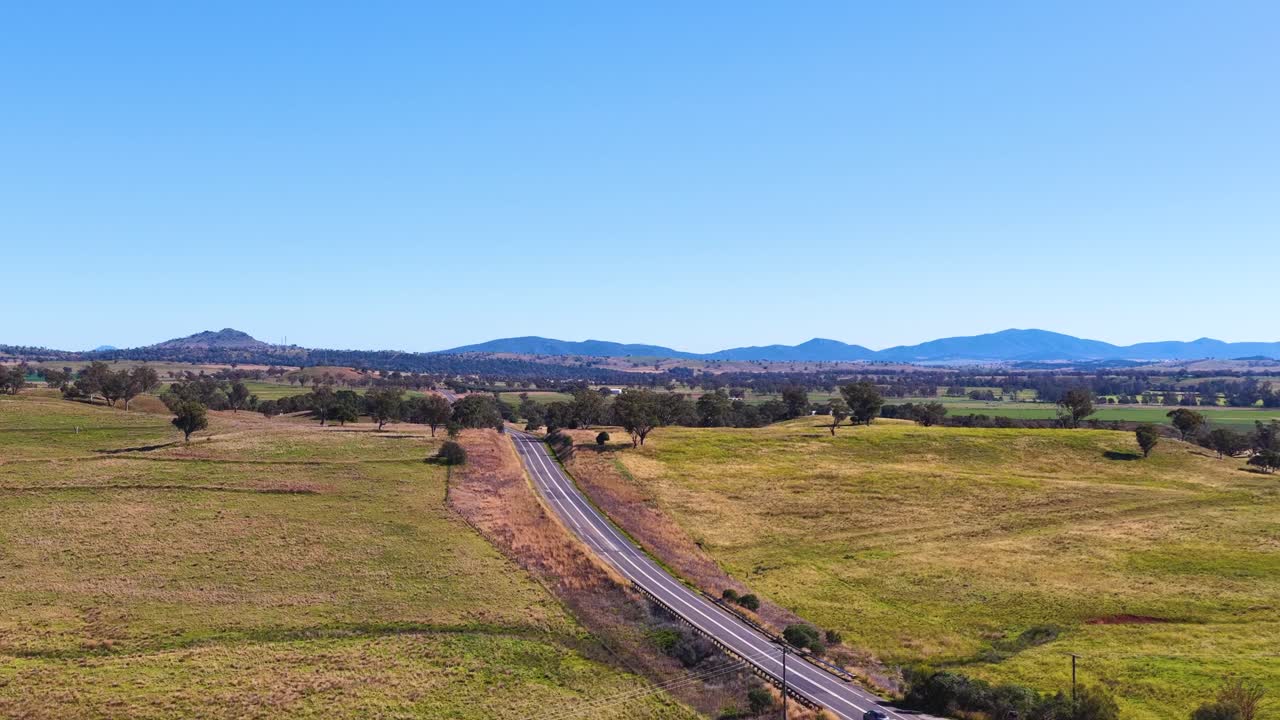 Drone footage glides above a winding rural road bordered by grassy fields, scattered trees, and distant hills under bright, clear daylight