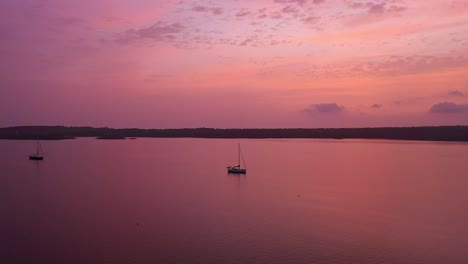 aerial view of two boats in the ocean in menorca spain during a pink, purple, orange sunrise, tracking wide shot