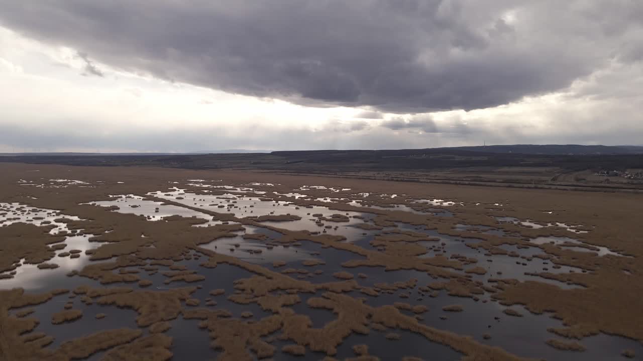 An aerial view of a vast wetland with patches of water reflecting the moody sky above. Dark clouds create a dramatic atmosphere over the natural landscape.