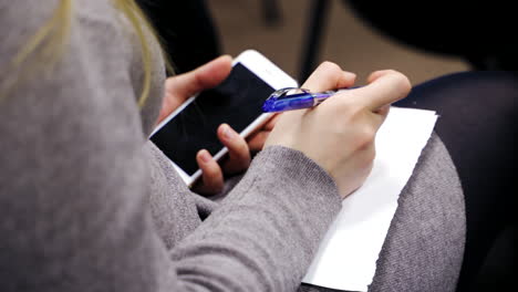 Woman is sitting and writing with a pen on a sheet of paper. Business woman holding phone and writes important information on paper during the conference indoors.