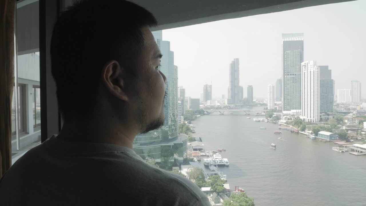 hombre asiático relajándose mirando por la gran ventana de la habitación del hotel con vista a la ciudad de bangkok en la orilla del río en vacaciones