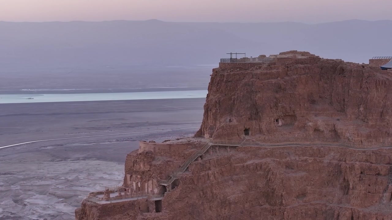 Veduta dall'alto e distante che cattura le aspre strutture dell'altopiano montuoso della fortezza di Masada contro un cielo crepuscolare viola e nebbioso, con la costa del Mar Morto che si estende sullo sfondo.