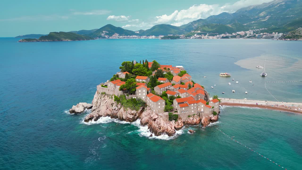 Sveti Stefan Island - Montenegro. Waves breaking on the rock. Famous and luxury travel destination. Boats in the bay. Turquoise water. Drone circling around the island