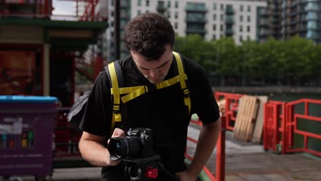 Kreisende-Aufnahme-Eines-Jungen-Kaukasiers,-Der-Seine-Kamera-Und-Sein-Stativ-Hält,-Während-Er-An-Einem-Sonnigen-Tag-Die-Straßen-Von-Canary-Wharf-In-London-Filmt