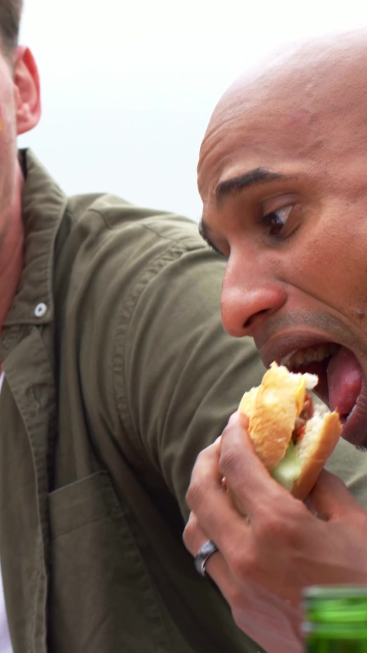 Vertical video: Talking bald man blue shirt replying to pal gesture at park table, biting sandwich