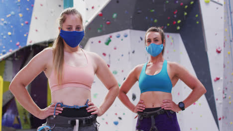 Porträt-Von-Zwei-Kaukasischen-Frauen-Mit-Gesichtsmasken,-Die-An-Einer-Indoor-Kletterwand-Stehen