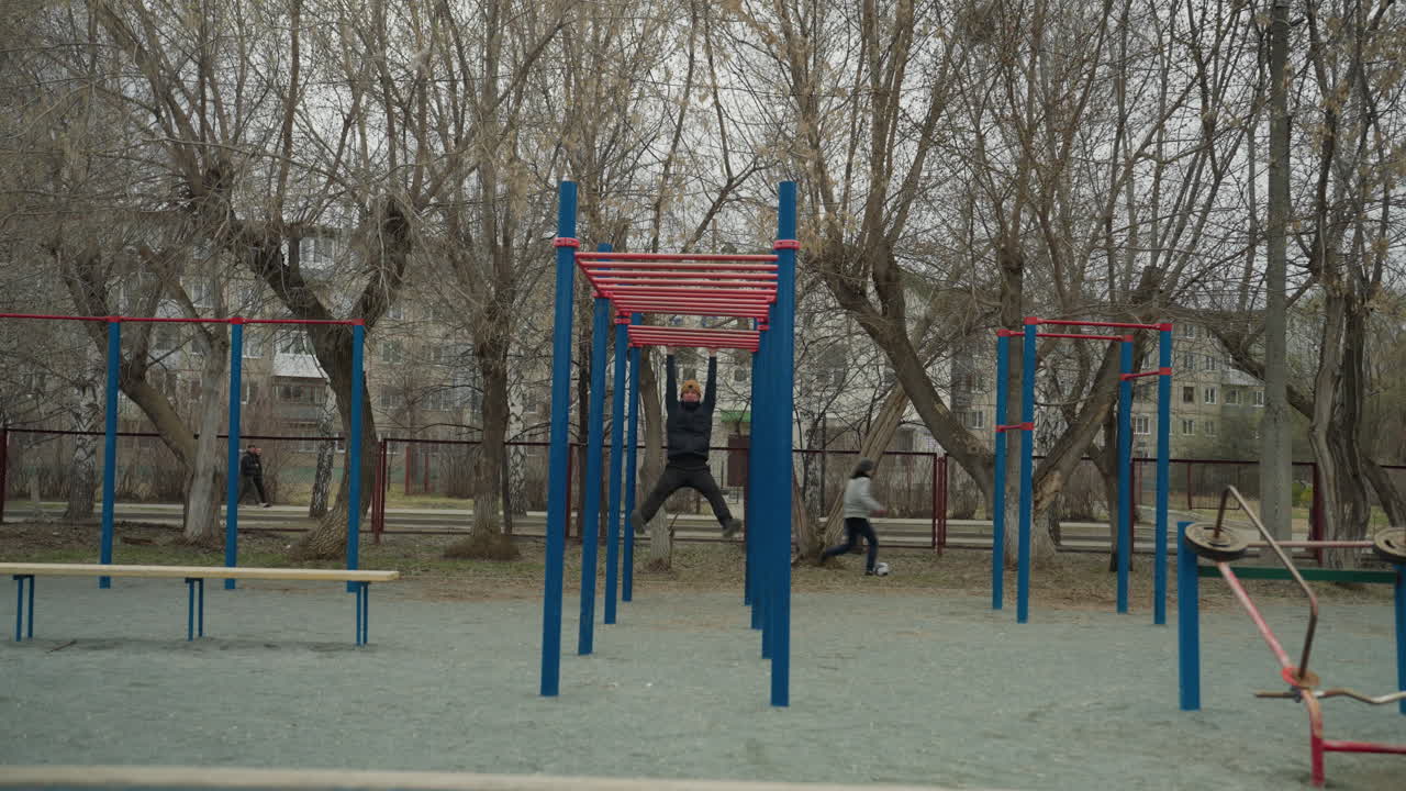 A coach jumps up to grab a red iron bar and begins to swing on it in an outdoor gym setup, in the background, someone is playing football, and cars are passing by with people walking