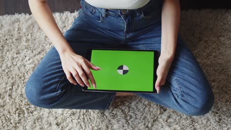 mujer en vaqueros se sienta en posición de loto en el suelo y utiliza tableta electrónica con pantalla verde, vista superior. maqueta. concepto de aparatos digitales