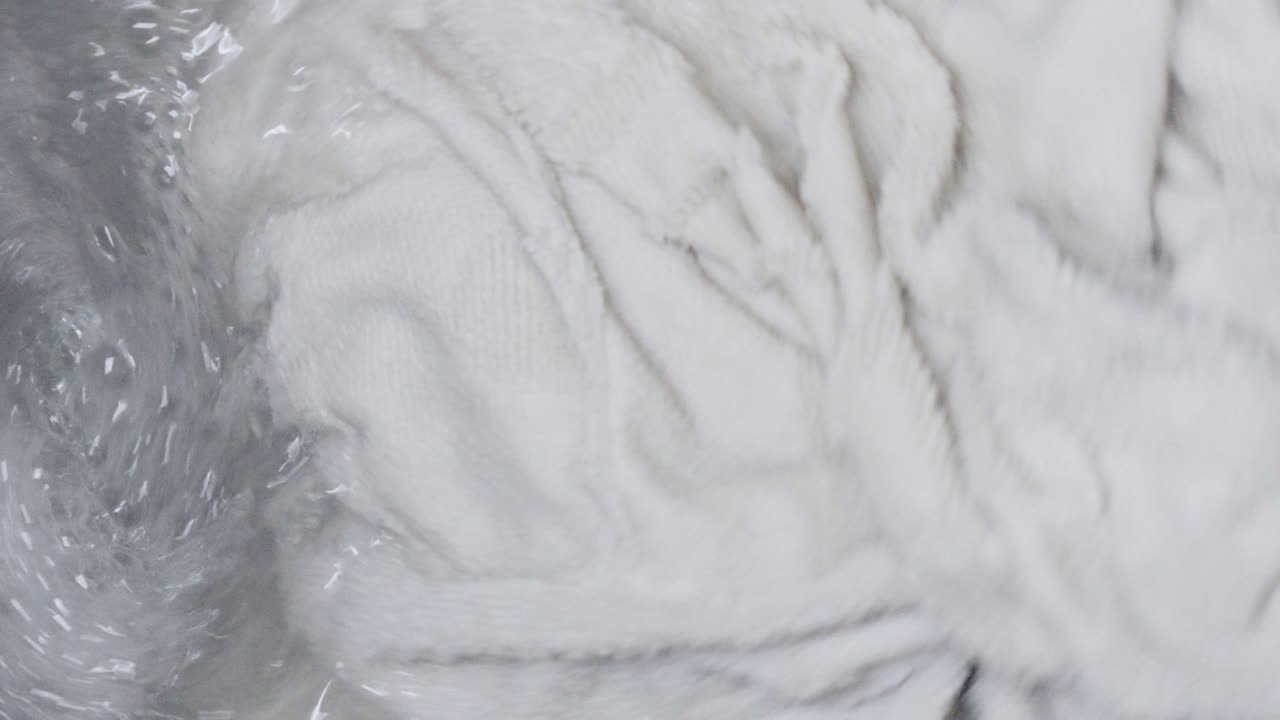 Primo piano di bucato bianco, principalmente asciugamani, che gira nel cestello di una lavatrice. L'acqua saponata vortica intorno ai vestiti bagnati durante un ciclo di lavaggio