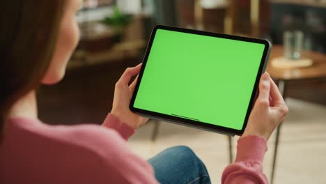 female holding tablet computer with green screen mock up display. woman relaxing at home, watching videos and reading social media posts on mobile device. close up over the shoulder footage.