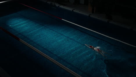 a woman swims freestyle in an indoor pool at night.