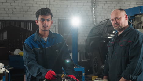 inventor in blue uniform stands near car engine in automotive workshop, illuminated by bright light, background features tools, equipment, and mechanical components