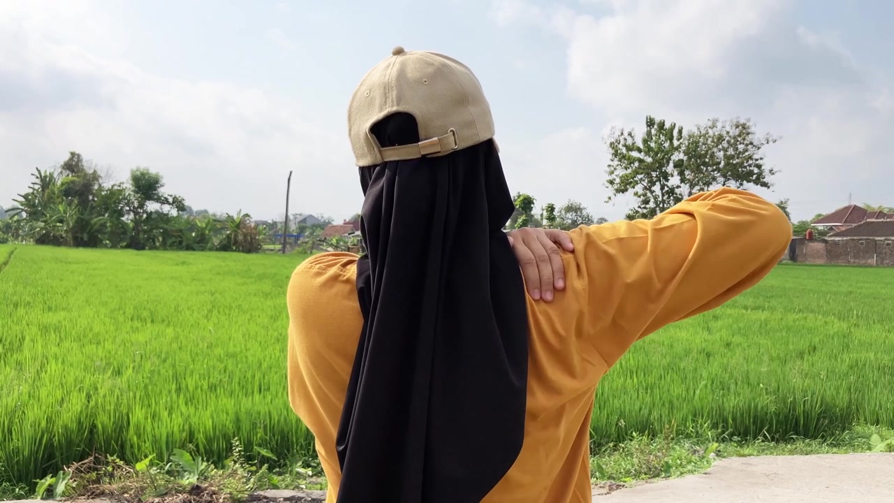 Pemandangan dari belakang dalam gerak lambat seorang wanita Muslim berhijab dan bertopi memegangi bahu kanannya kesakitan. Dia mengalami nyeri otot saat berdiri di luar ruangan di samping sawah hijau yang subur.