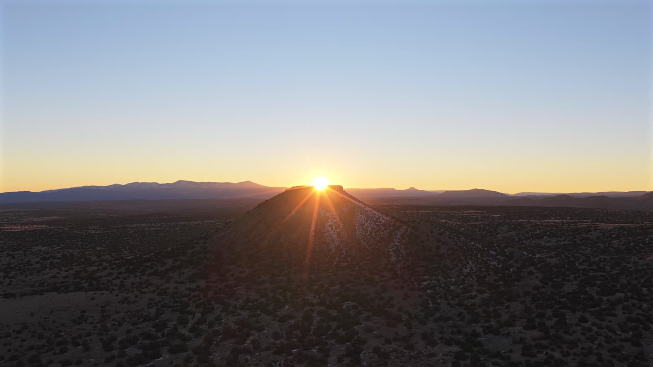 An aerial push toward a rugged desert mesa as the sun steadily breaks over the peak. This cinematic 4K transition features a brilliant starburst and golden light across a vast wilderness