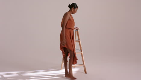on cue indian woman in rust-orange dress, circling wood ladder, sitting, adjusting hair in studio