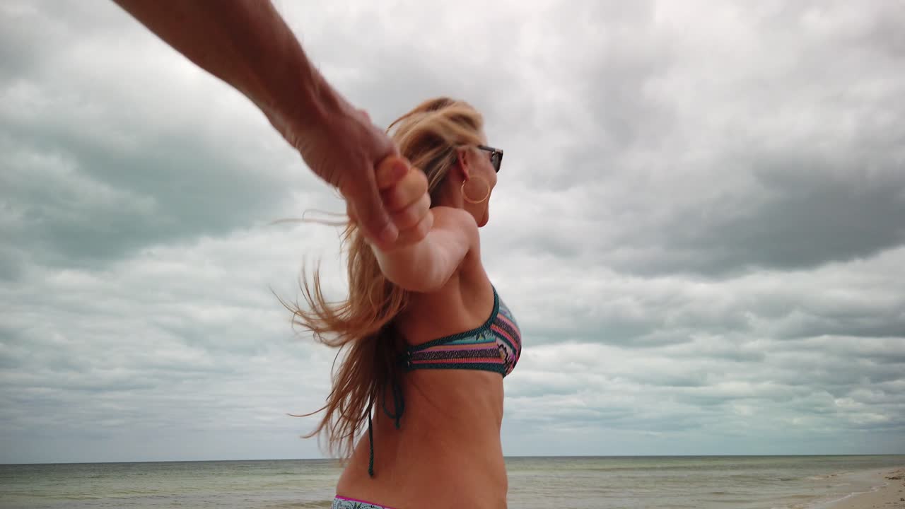 성숙한 여인은 해변에서 남편을 끌어당기는 나를 따라 슬로우 모션으로 비키니와 선글라스를 달린다