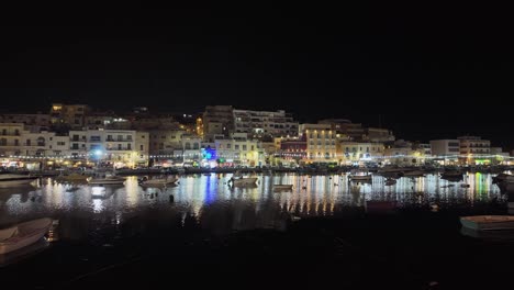 Vista-Nocturna-Del-Centro-De-La-Ciudad-De-Marsaskala-A-Través-De-La-Bahía-Mediterránea-Llena-De-Barcos-Con-El-Paseo-Marítimo-Iluminado-Y-Restaurantes-Al-Fondo-En-Malta