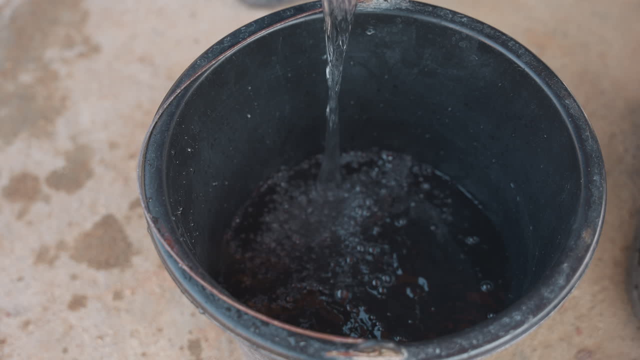 Primo piano di un secchio che si riempie d'acqua su un pavimento di cemento, getto costante che forma increspature e morbidi schizzi, secchio di plastica scuro che mostra sottili riflessi, luce naturale ambientale, la scena suggerisce la preparazione dell'officina