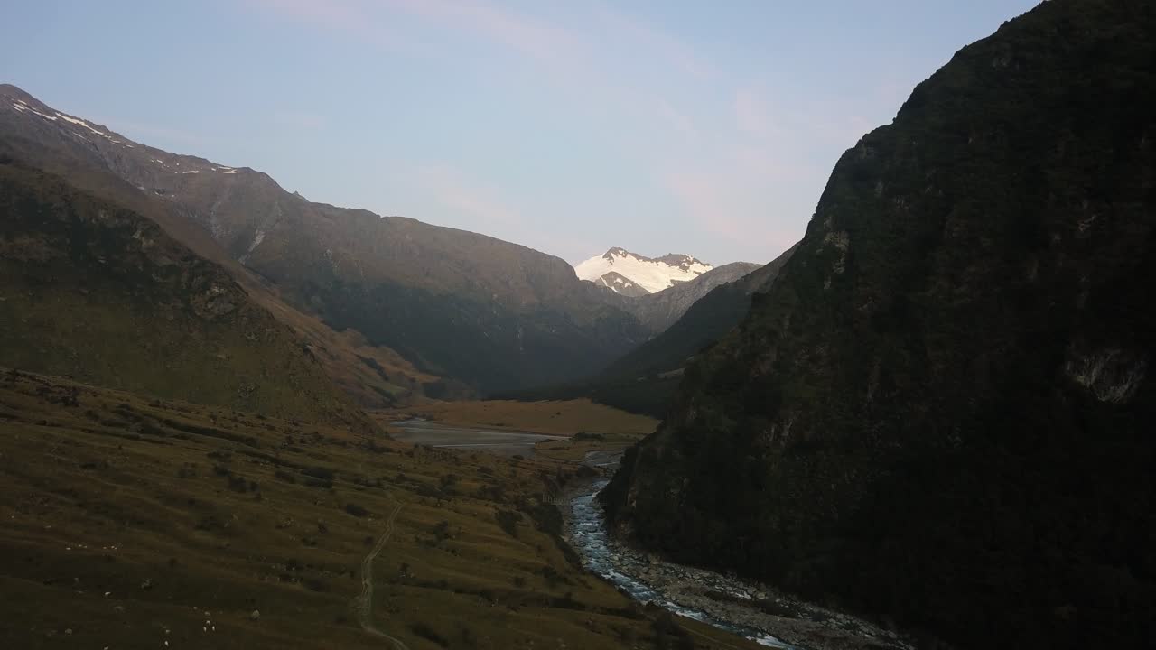 Moving forward aerial shot through a dark winding river valley with an illuminated snow capped mountain glacier in the far distance