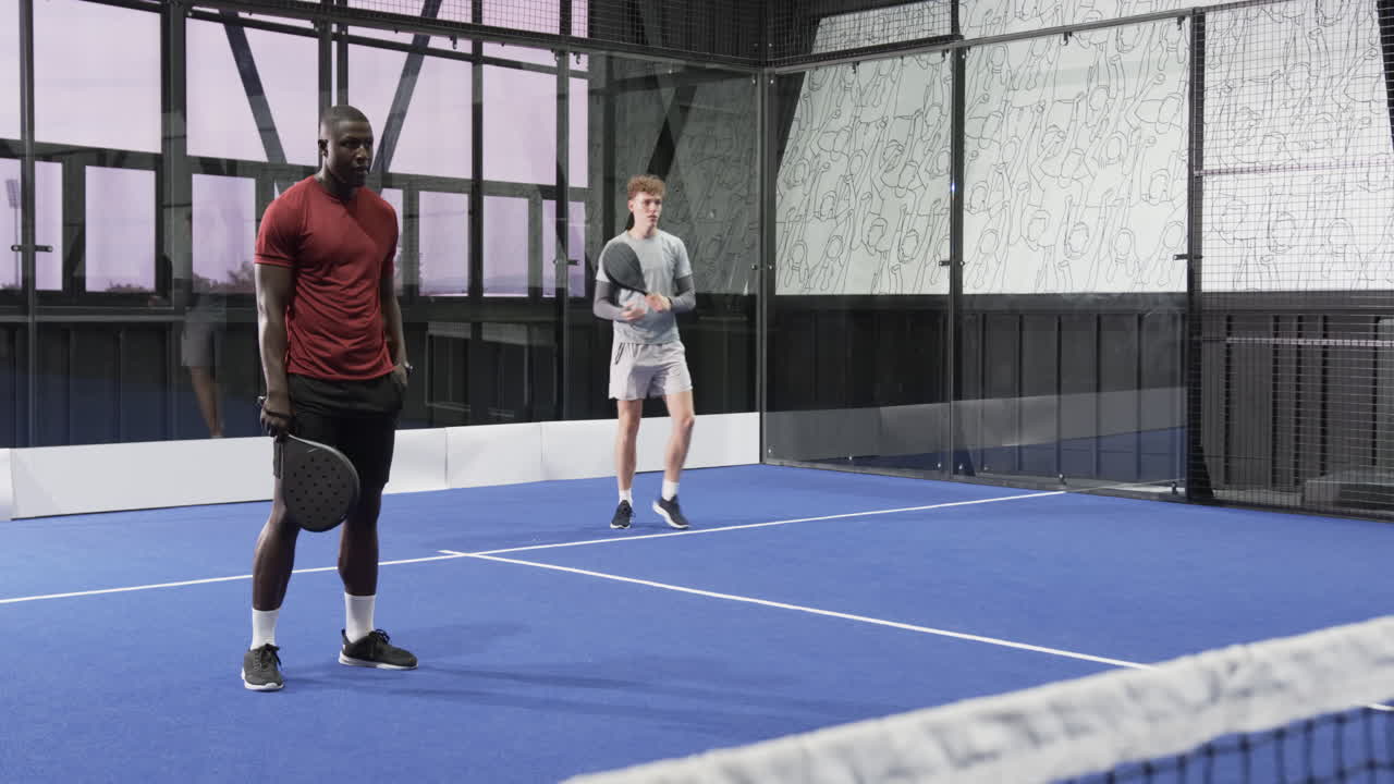 Two diverse men playing padel tennis on indoor court, focused and ready for action