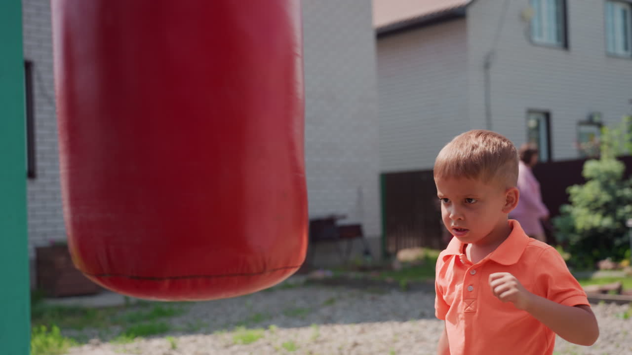 Male Youth In Orange Shirt Trains With Punching Bag During Summer Afternoon, Young Male Athlete Wearing Orange Polo Actively Practicing Boxing On Red Punching Bag In Outdoor Garden Setting