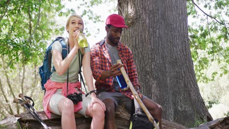 happy diverse couple with backpacks sitting and drinking water in park, slow motion