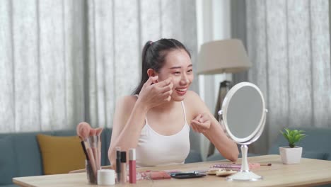 hermosa joven asiática se mira en el espejo aplicando crema facial y sonríe mientras se maquilla en casa