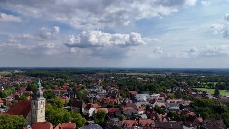 aerial view of a picturesque german town with red-tiled roofs, historic church tower and lush green surroundings under partly cloudy summer sky. forward flight. wide shot. wind turbine in back