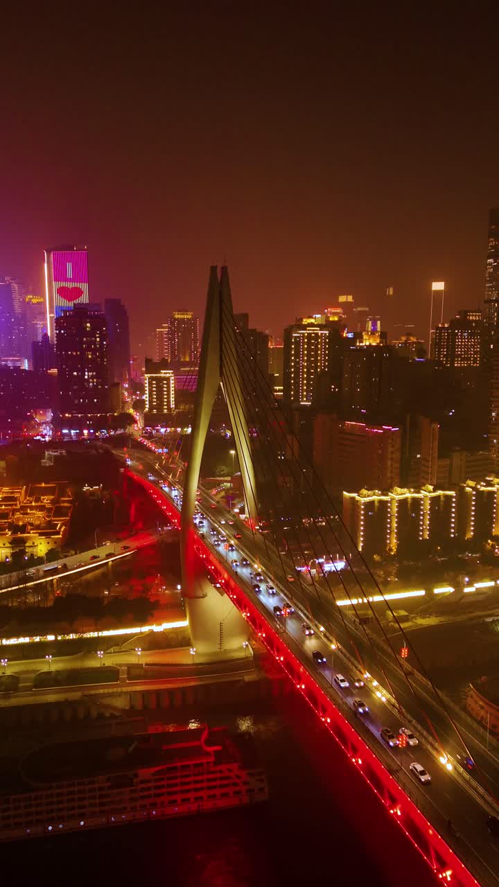 Vertical aerial nighttime view of the Dongshuimen Bridge in Chongqing, China, illuminated with vibrant red lights. Traffic flows across the bridge with skyscrapers and glowing city lights