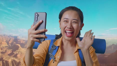 close up of asian female hiker with mountaineering backpack waving her hand and pointing back while having a video call on smartphone while traveling at the top of mountain