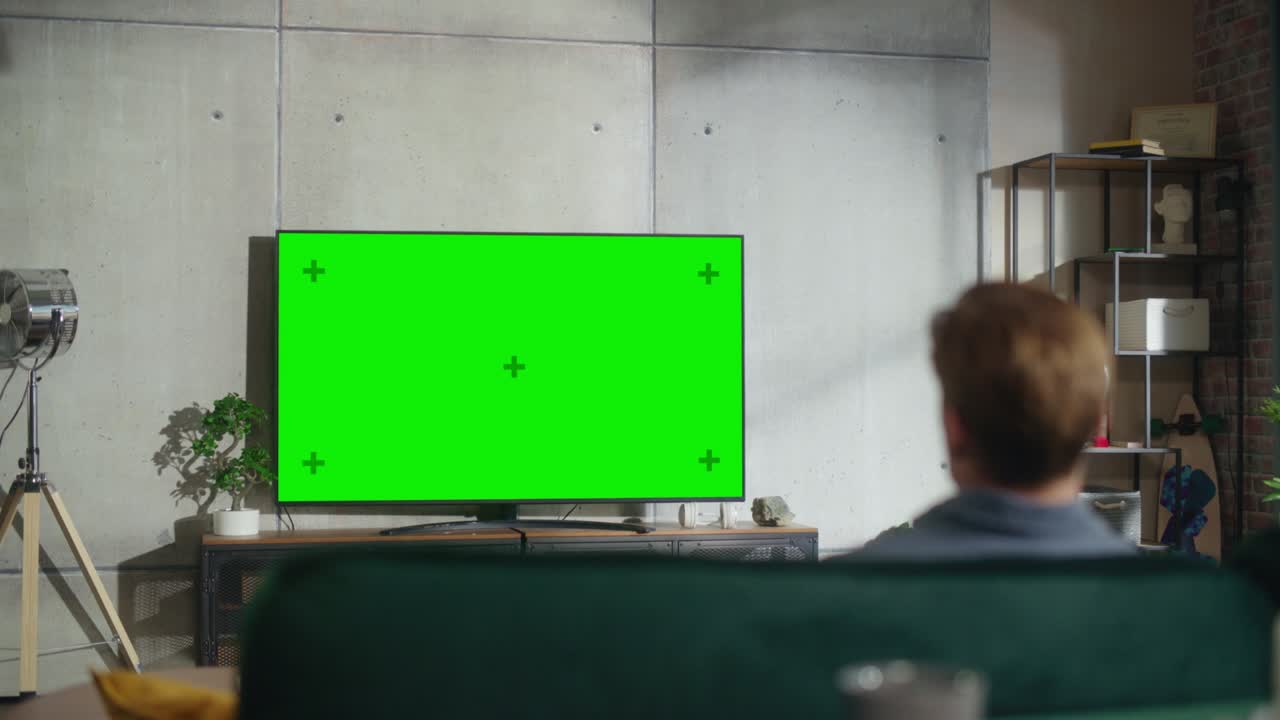 젊은 예쁜 남자가 집에서 시간을 보내고, 소파에 앉아서 초록색 화면으로 tv를 켜고, 세련된 로프트 아파트에서 모의 디스플레이를합니다.