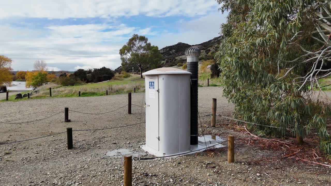 Aerial camera pans across a small public restroom in a gravel parking area, surrounded by trees, hills, and open countryside under daylight
