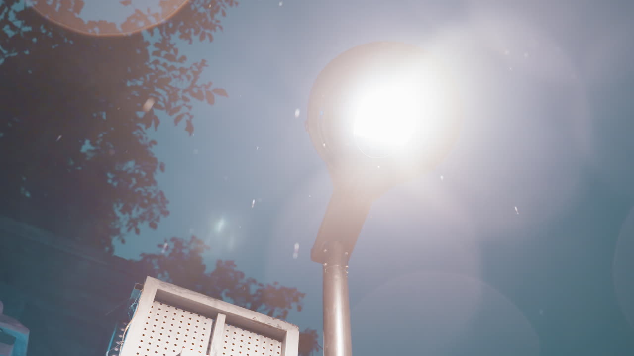 Sinal retrô de poste de luz brilhante à noite. Poste elevado emite um brilho quente e bokeh contra um céu azul-petróleo frio, galhos de árvores em silhueta emoldurando a cena. Letreiro vintage com luzes pontilhadas sugere um ambiente urbano vibrante.