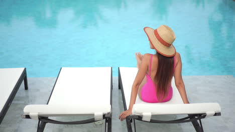 back view of young woman with large hat and swimsuit on deckchair by pool