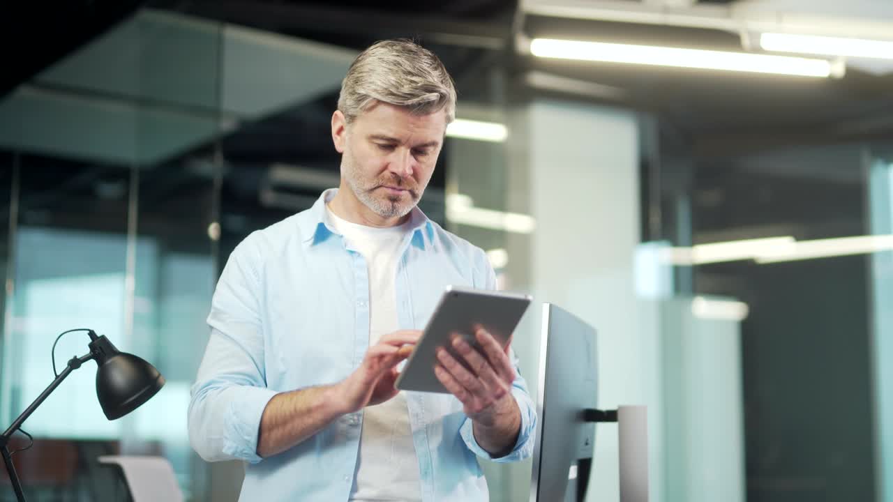 зрелый бизнесмен использует цифровой планшет, стоящий в современном офисе в помещении. седоголовый старший мужской сотрудник использует просмотр смартфона.