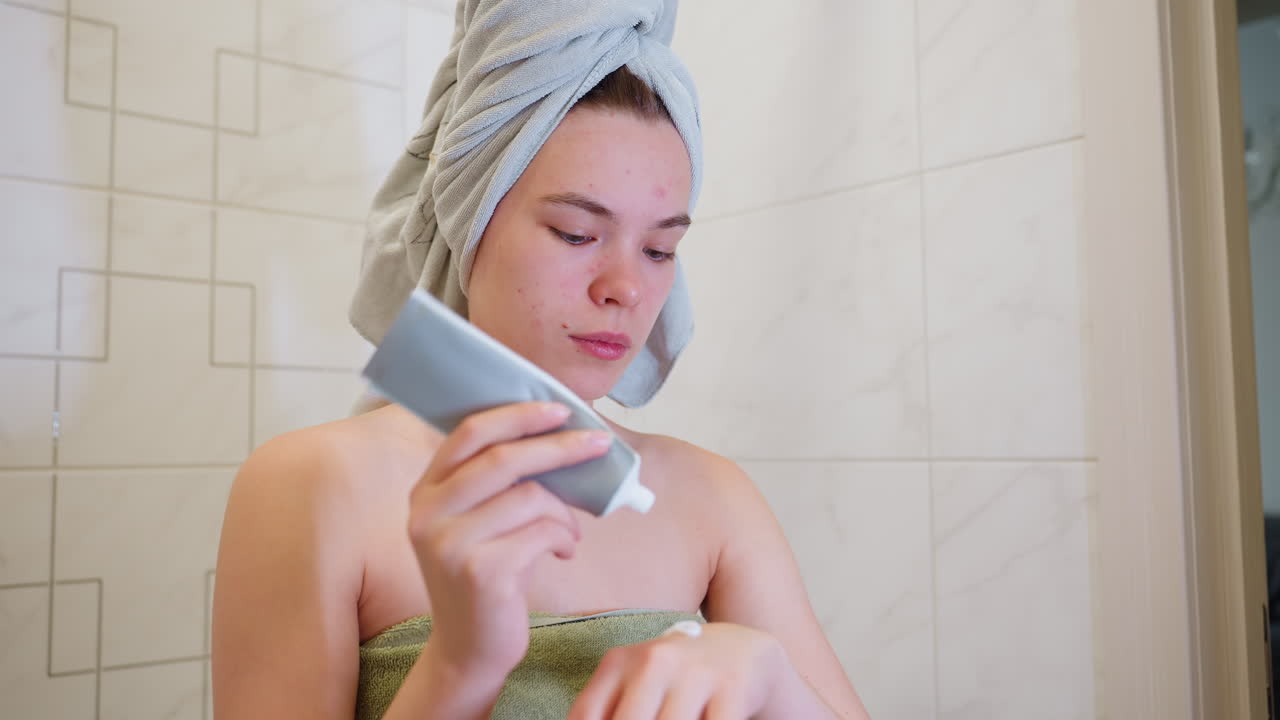 Close-up view of woman applying cream on her hand, focusing on skincare routine, she is in bathroom with towel wrapped around her hair, carefully moisturizing her skin after shower
