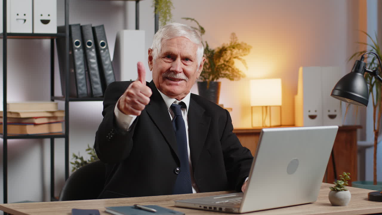 Happy caucasian elderly businessman using laptop and showing thumbs up at camera in home office