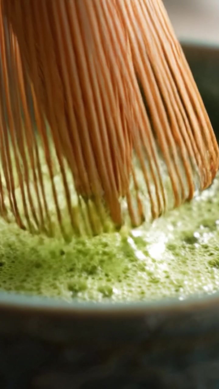 Una vista ravvicinata di un frullino di bambù che prepara il tè Matcha, mostrando un ricco colore verde e bolle in una ciotola di ceramica durante una tradizionale cerimonia del tè giapponese.