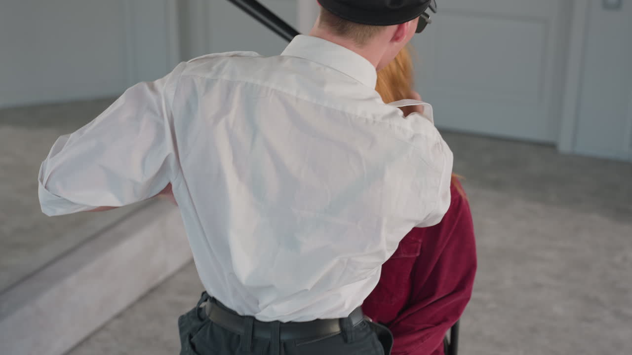 Back view of man in white shirt leaning toward woman in red shirt sitting on chair, her face partially hidden by his body, suggesting intimate playful interaction in minimal modern interior