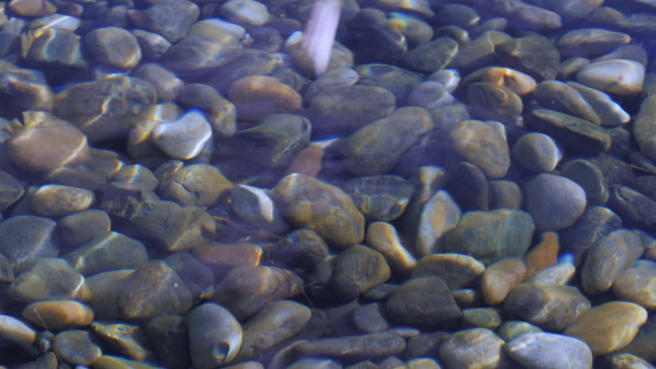 Rock splashing on reflective smooth clear water surface with pebbles under, slow motion.