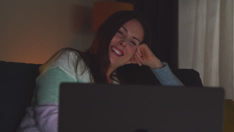 smiling woman sitting on sofa at home at night streaming or watching movie or show on laptop 1