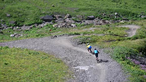 two riders are going downhill with their bikes on a dirt and gravel path next to meadows in the swiss alps, obwalden, drone view