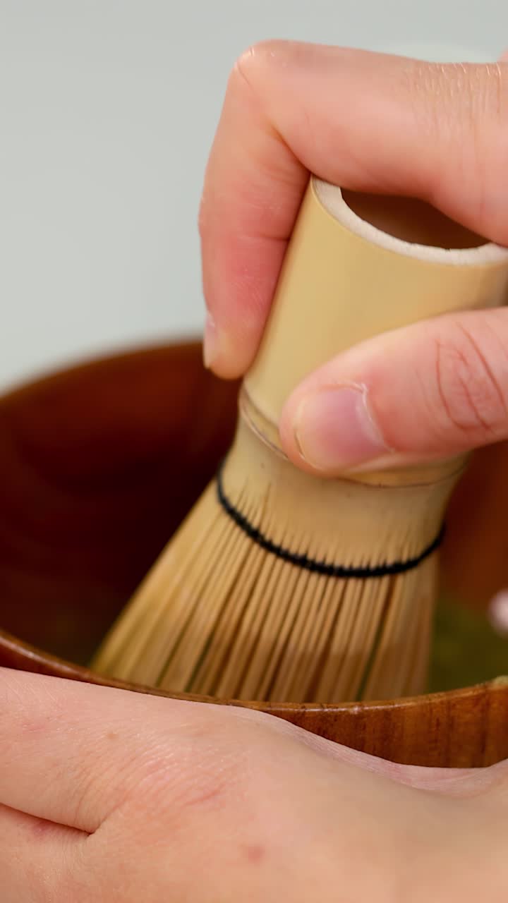 Primo piano di mani che sbattono tè verde matcha in una ciotola di legno sotto una morbida illuminazione da studio.