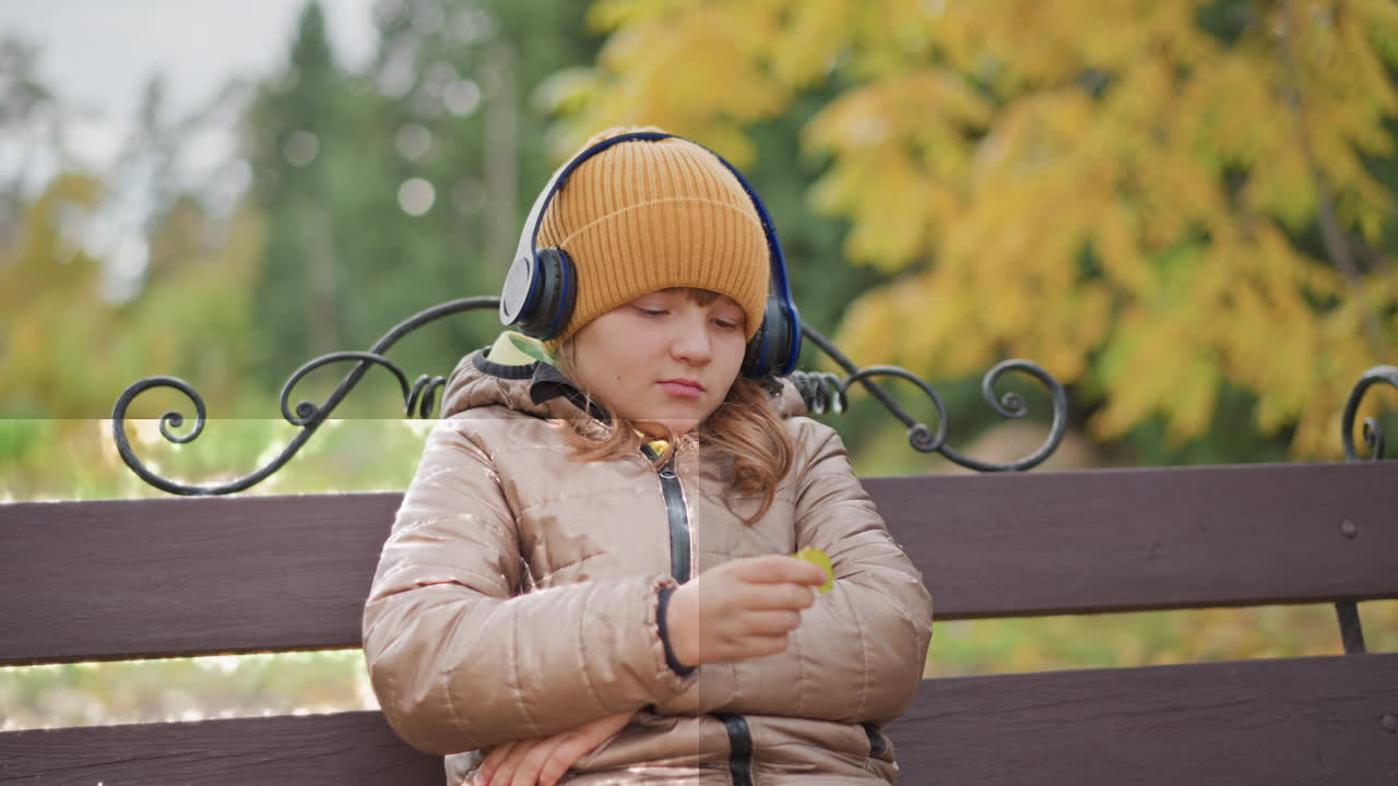 young girl in mustard beanie and headphones sits on park bench under golden autumn trees, thoughtfully squeezing yellow leaf between fingers while immersed in music, crisp air swirling fallen leaves
