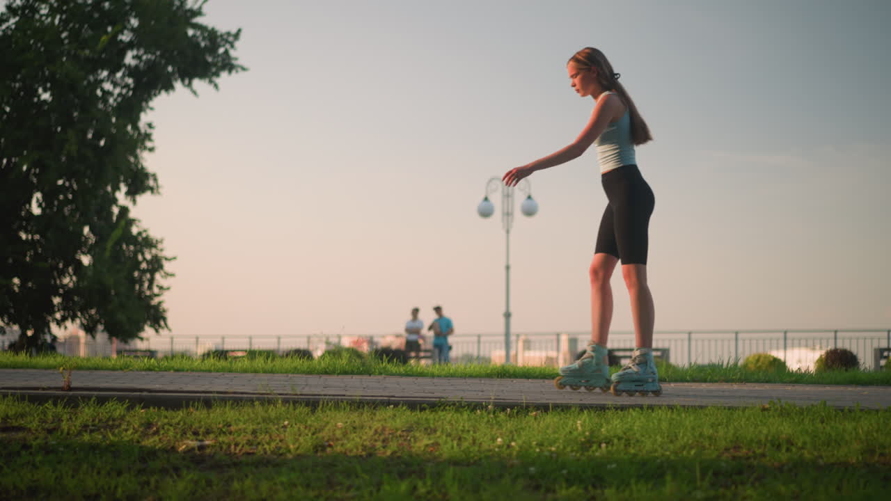 은 공원에서 야외에서 롤러 스케이팅하는 젊은 여성, 균형을 위해 손을 흔들고, 배경에 흐릿한 도시 풍경, 마천루, 울창한 초록색, 멀리서 두 사람이 둘러싸여 있습니다.