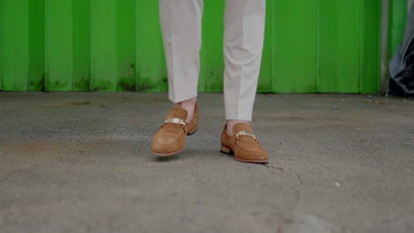 Man-wearing-beige-pants-and-brown-loafers-walking-on-a-pavement-in-front-of-a-green-wall