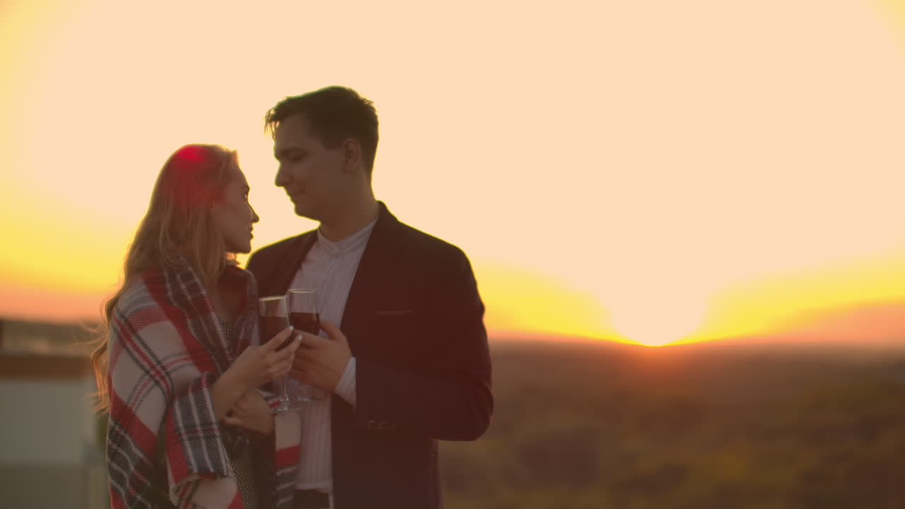 Giovane coppia sposata sul tetto che si abbraccia e beve vino rosso dai bicchieri, in piedi e vestita a quadri, ammirando il bellissimo tramonto sulla città.