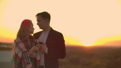 una joven pareja casada en el techo abrazándose y bebiendo vino rojo de vasos de pie vestidos de a cuadros y admirando la hermosa puesta de sol sobre la ciudad...