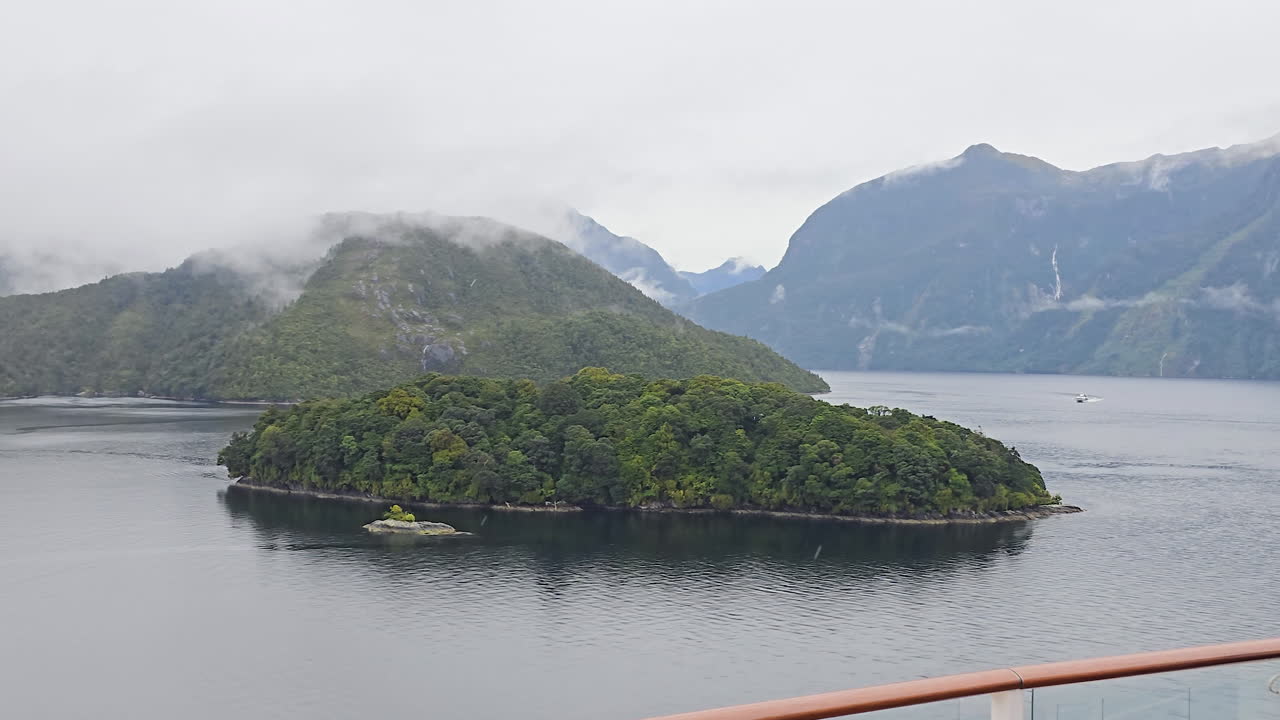 Viewing scenic landscape of Milford Sound in New Zealand exploring the fjord from cruise ship, showing small forested island with mist shrouded mountains and waterfalls
