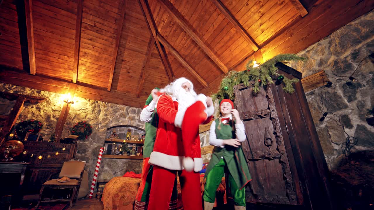Santa Claus is wearing his costume in specially decorated room in his bed. Boy and girl elves are helping him to wear red jacket and a hat. Christmas spirit concept close up video. Christmas eve. New Year is coming. Aerial view. Video from below.